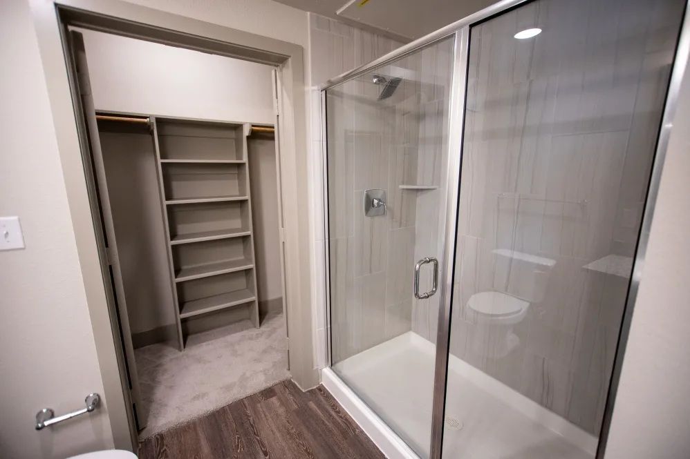 Bathroom with walk-in closet, glass shower, toilet, shelving. Light wood floors, light grey walls.