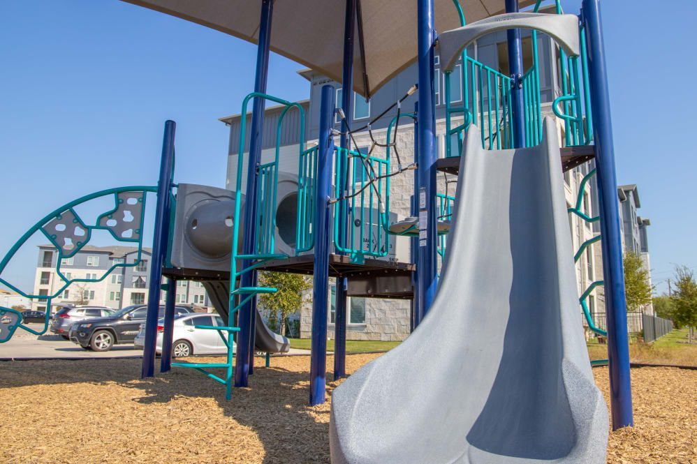 Playground with blue and gray structures, slide, and climbing elements on wood chips. Apartments in the background.