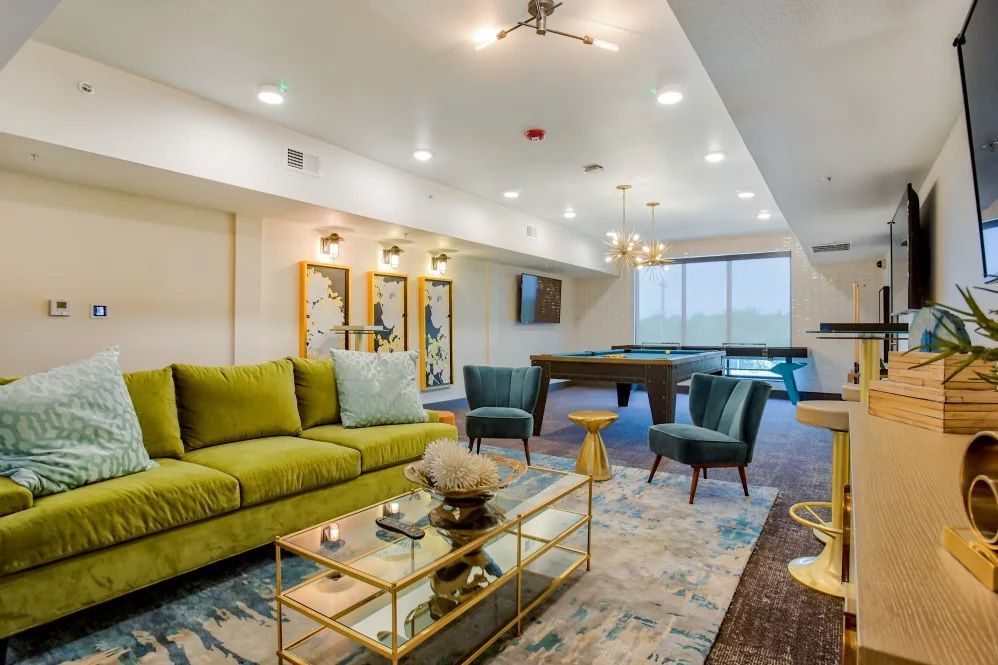 Lounge area with green couch, pool table, and multiple chairs. Well lit, modern interior with a light blue rug.