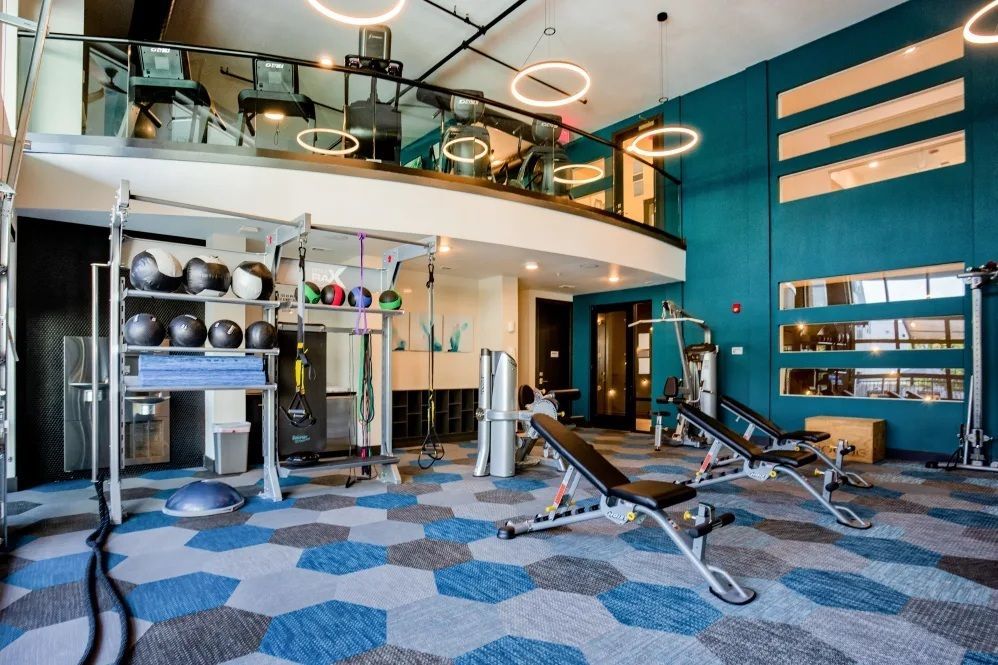 Gym with exercise equipment, including weights and benches, against teal walls and a second-floor mezzanine.