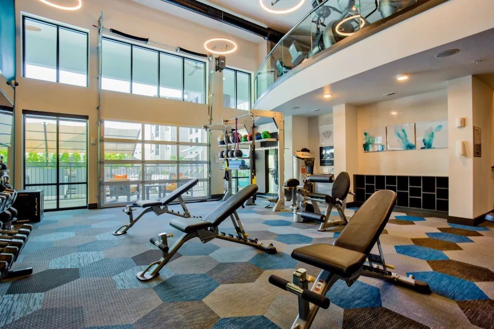 Gym with weight benches, dumbbells, and exercise equipment. Large windows and a second-floor walkway. Hexagonal patterned flooring.