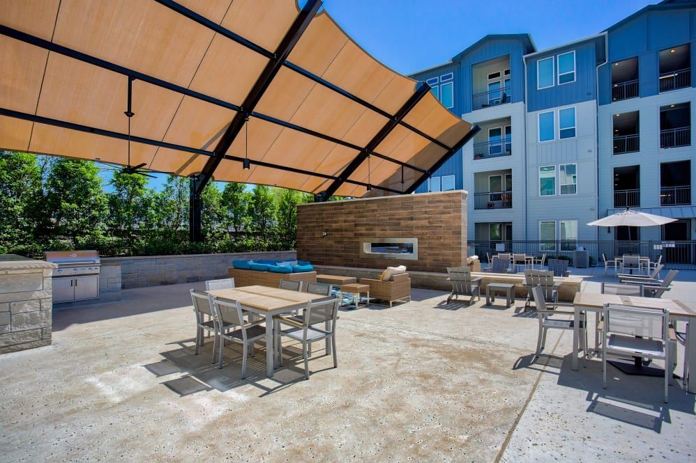 Outdoor patio with tables, grill, hot tub, fireplace, and shade structure. Modern apartment building in the background.