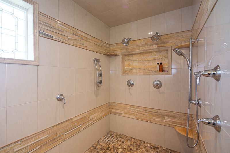 Bathroom with beige tile, stone floor, multiple shower heads, built-in bench and shelf.