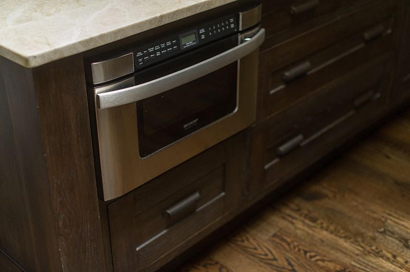 Built-in stainless steel microwave in dark wood kitchen cabinetry with stone countertop and wood floor.