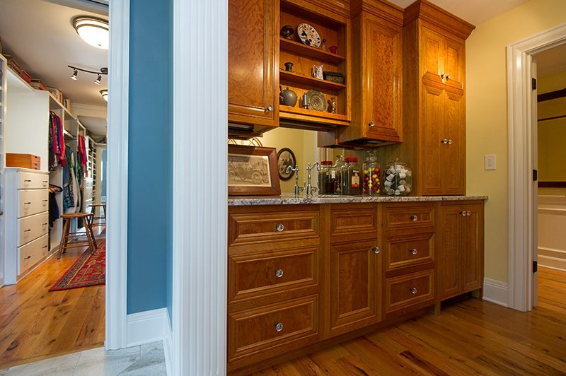 Wooden cabinetry in a room with a walk-in closet. Blonde wood floor and colorful clothes inside the closet.