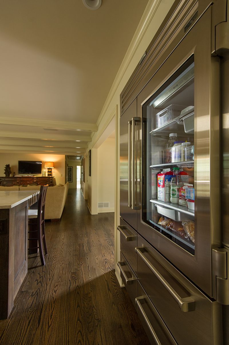Stainless steel refrigerator with glass door in a kitchen, showcasing interior contents; open layout with hardwood floors.