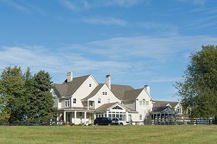 Large, light-colored house with multiple gabled roofs, set in a grassy field, under a blue sky.