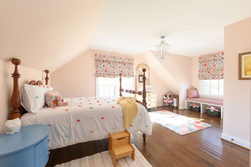 Pink girl's bedroom with a bed, floral window shades, and a bench, on hardwood floors.