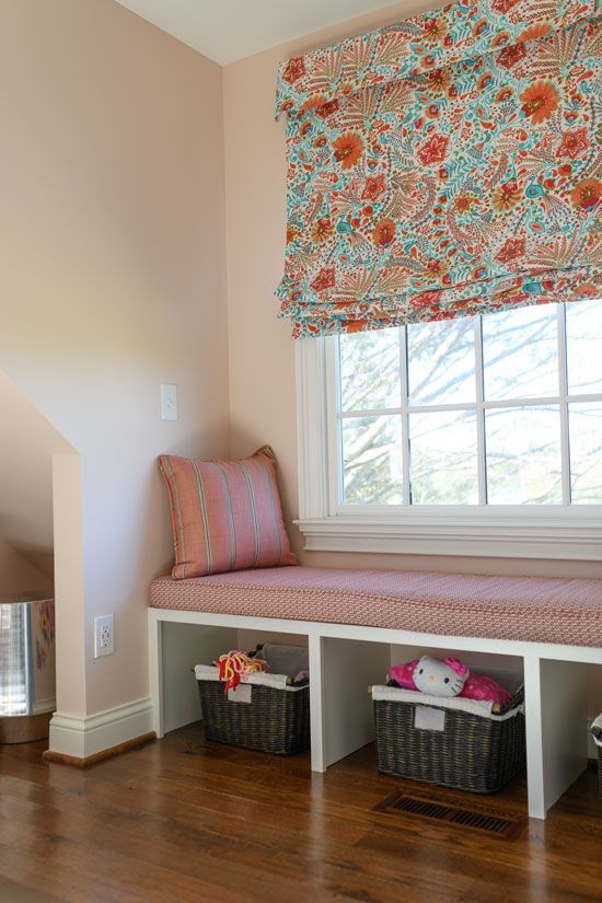 Cozy window seat with patterned cushion, storage baskets below, floral window shade, and pink walls.