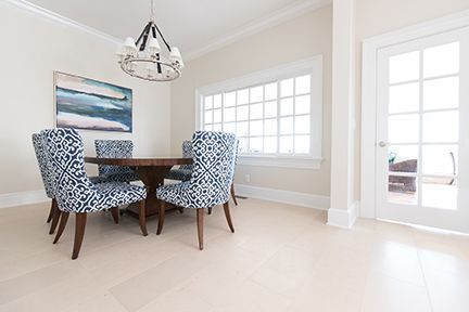 Dining room with round wooden table, blue patterned chairs, painting, large window, and chandelier.