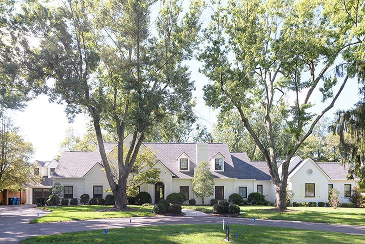 White brick house with trees, driveway, and blue trash cans.