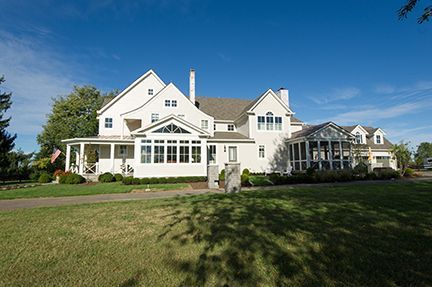 White house on a grassy lawn under a bright blue sky. A covered porch and glass sunroom are visible.