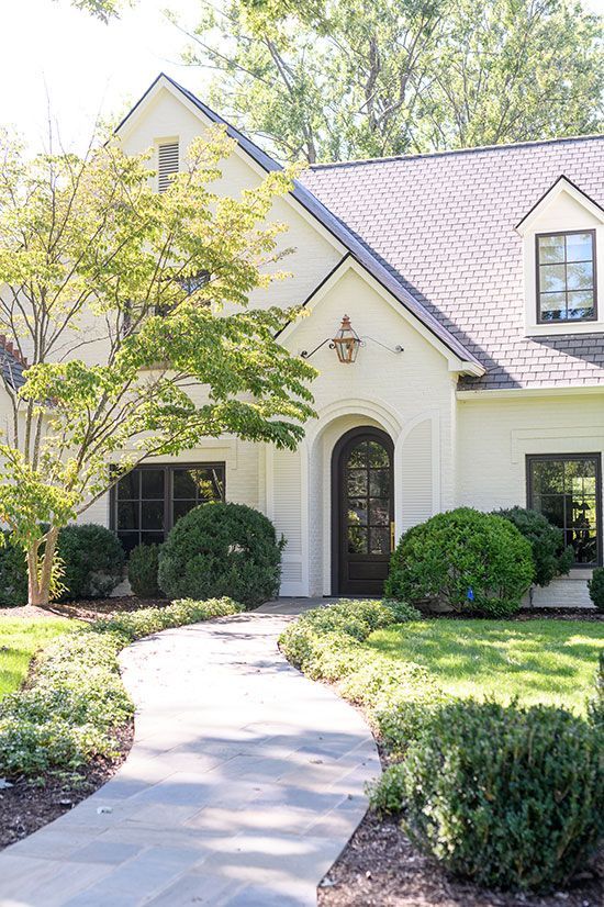 White house with a gray roof, dark arched door, and a winding stone path. Green shrubs and trees frame the entrance.