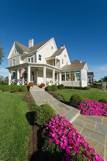 Beige house with porch, flag, stone walkway lined with pink flowers, green lawn under blue sky.