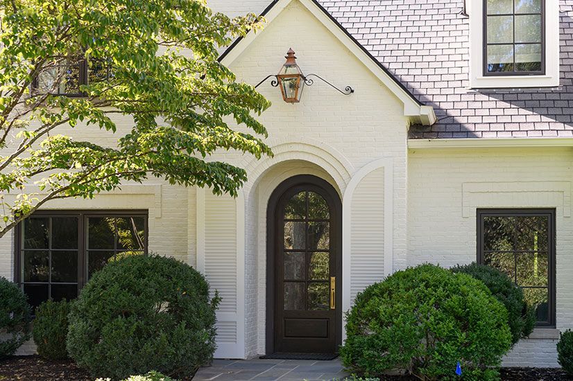 White brick home with dark brown door and windows; lush greenery frames the entrance.