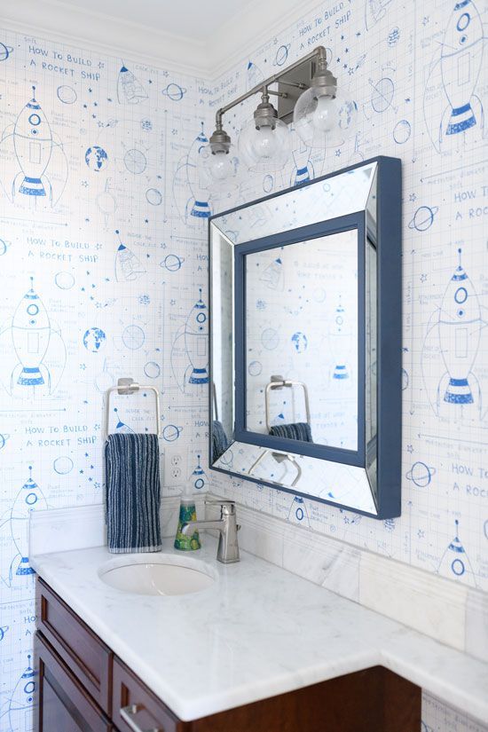 Bathroom with blue-framed mirror, white countertop, blue patterned wallpaper, silver fixtures, and a dark wood cabinet.
