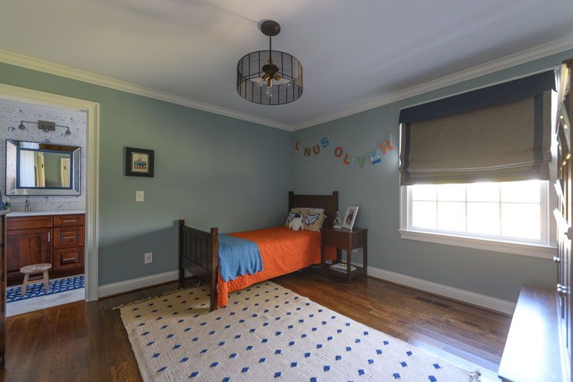 Child's bedroom with a bed, rug, and adjacent bathroom; blue walls, hardwood floor, and a modern light fixture.