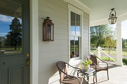 A porch with two chairs, a small table, a lantern, and a door, surrounded by a yard.