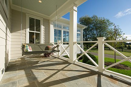 Porch with swing, white columns, gray siding, and stone floor. Sunlight casts shadows.
