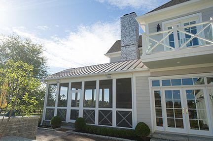 Exterior of a light-colored house with a screened porch and balcony. Bright sky, chimney, and landscaping visible.