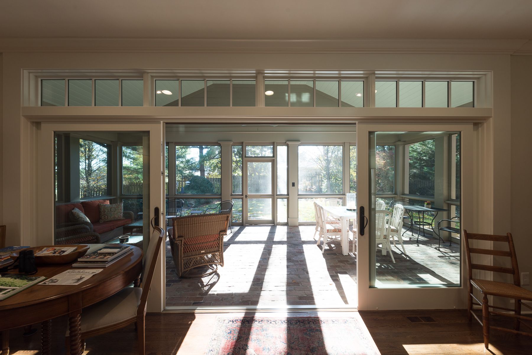 Interior view of a sunroom with open glass doors casting shadows on a dining table and chairs.