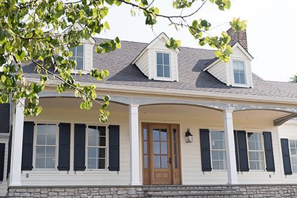 White house with a stone foundation, blue shutters, and a wooden door. Trees in the foreground, sunny day.