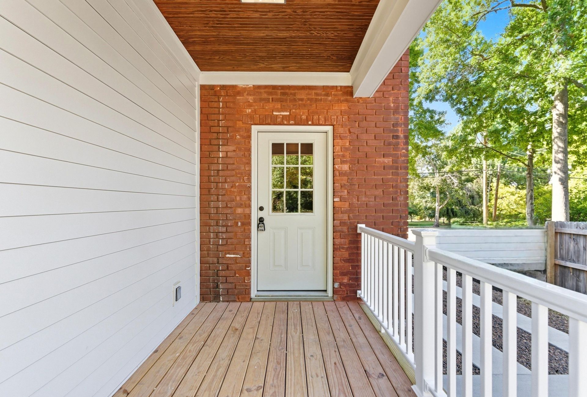 Front porch with a white door, brick wall, wooden deck, and white railing.