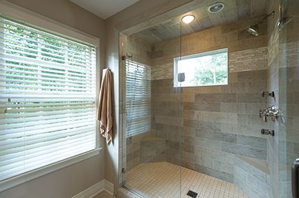 Bathroom shower with glass door, tile walls and floor, window with blinds, and a bench seat.