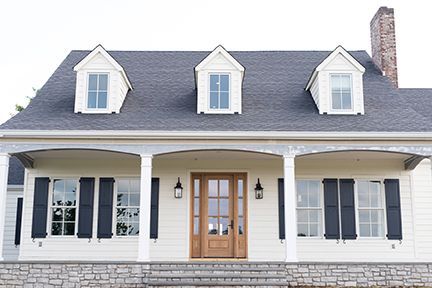 Cream house with a porch, black shutters, three dormer windows, and a brown door.
