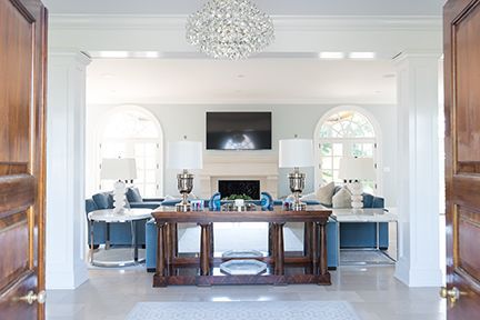 A bright living room with blue couches, a fireplace, and a crystal chandelier, viewed through open wooden doors.