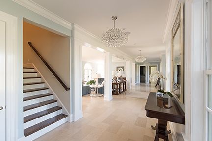 Elegant, light-filled hallway with chandelier, staircase, and dark wood table. Bright, spacious interior.