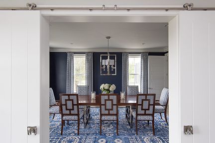Dining room with dark blue walls, long wooden table, ornate chairs, blue patterned rug, viewed through sliding white doors.