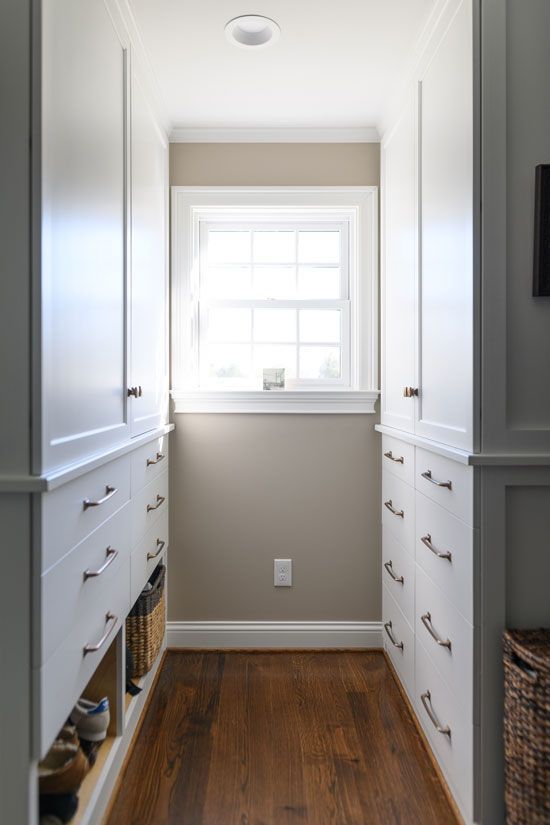 Narrow walk-in closet with white cabinets, drawers, and a window at the end. Wooden floor, beige wall.