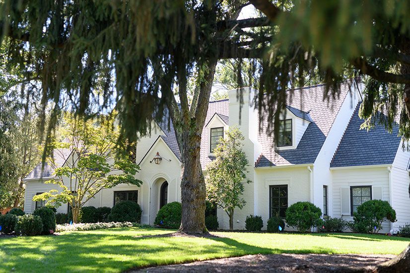 White house with black-trimmed windows, dark roof, and manicured lawn, framed by large trees.