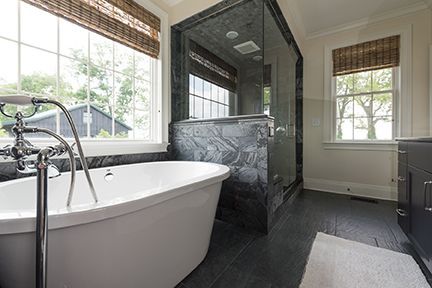 Bathroom with a white tub, glass shower, and two windows with bamboo shades. Black floor and walls.