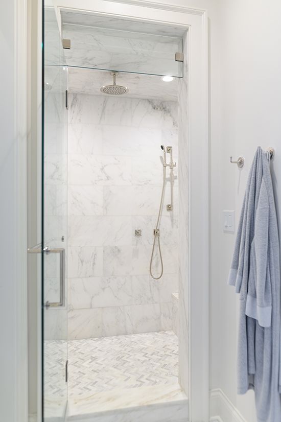 Narrow walk-in shower with white marble walls, a glass door, and a grey hexagon tile floor. A robe hangs nearby.