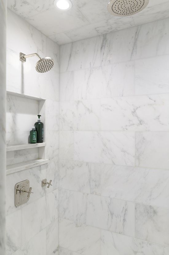 White marble tiled shower with two showerheads, shelves holding shampoo and body wash.