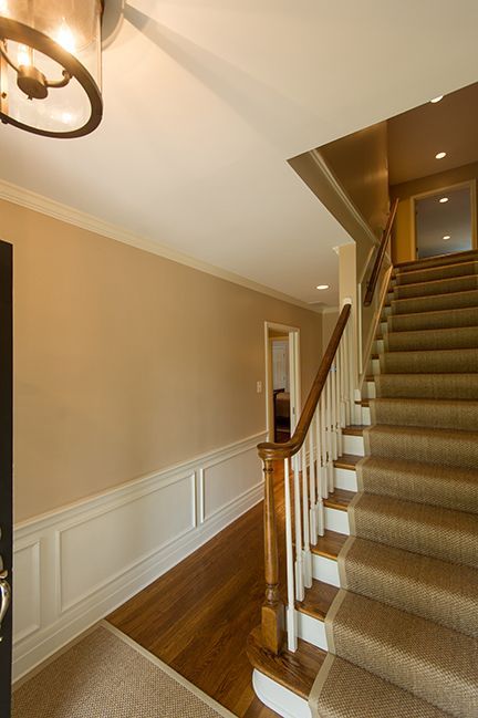 Hallway with stairs. Beige walls, white trim, brown handrail, carpeted steps, and wood flooring.