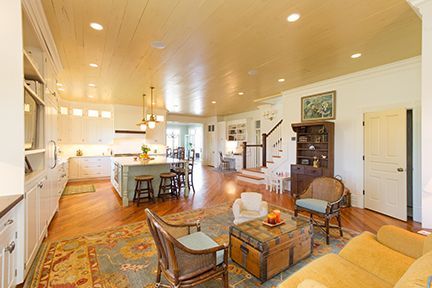 Open-concept living space with kitchen, living area, and stairs. Hardwood floors, white cabinets, blue rug, and wicker furniture.
