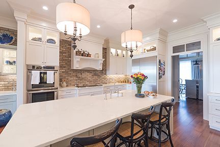 Elegant white kitchen with a large island, brick backsplash, and light fixtures.