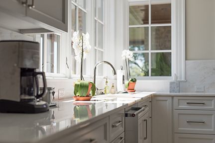 Bright kitchen with white cabinets, countertop, and windows; coffee maker, faucet, orchid plant, and sunlight.