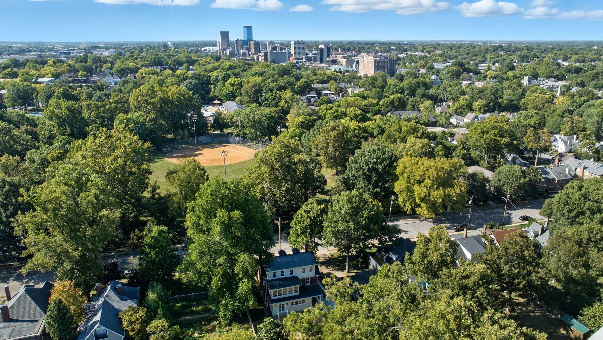 Aerial view of a city park with trees, a baseball field, and buildings in the distance under a blue sky.