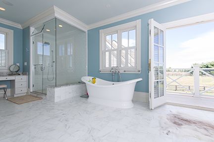 Bright blue bathroom with white marble flooring, glass shower, and a freestanding bathtub next to open French doors.