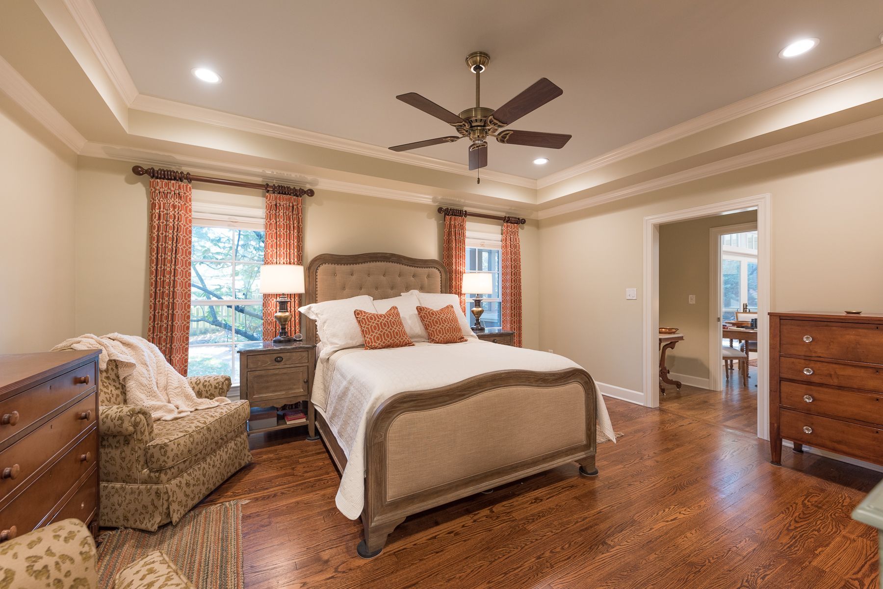 Bedroom with bed, wooden floor, beige walls, and ceiling fan. Two windows with red curtains.
