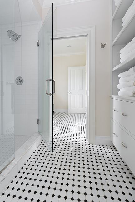 A white bathroom with black and white tiled floor, glass shower, open door, and shelves of towels.