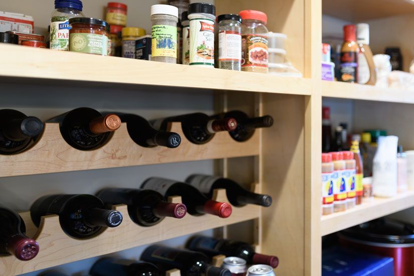 Wine bottles in a wooden rack below a shelf of various spice jars in a pantry.