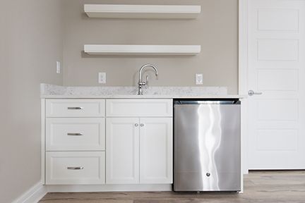 A white wet bar with a stainless steel mini-fridge, sink, and floating shelves against a beige wall.
