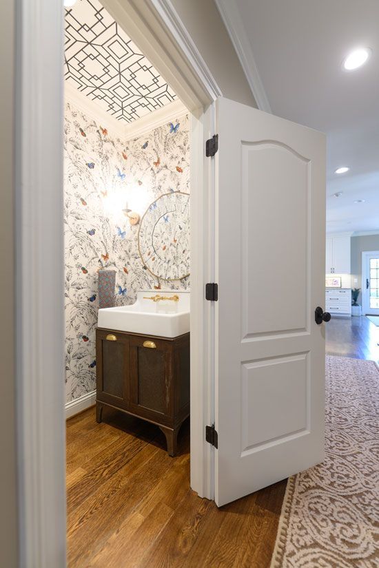 White door ajar to a powder room with patterned wallpaper and wooden vanity.