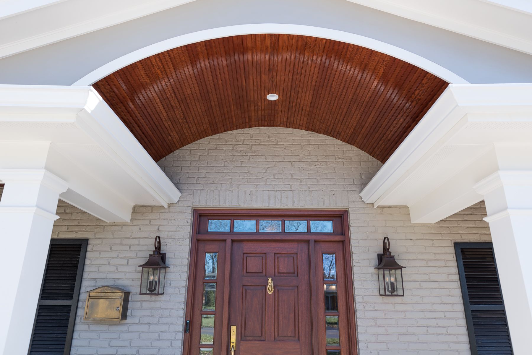 Front door with arched wood ceiling and white columns. Brown door, brick wall, and lanterns.