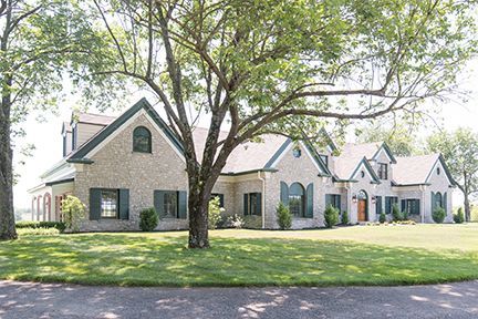 Large, light-colored brick house with green shutters and roof, set on a grassy lawn under a tree.